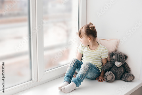 A little girl sits on a window sill and looks out the window into the street. It's a sad mood. Illness or self-isolation.