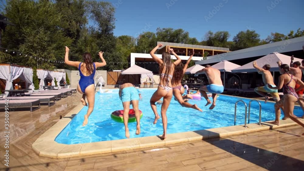 Happy friends jumping and splashing in swimming pool with inflatable floats in luxury resort. Young people in swimwear having party in private holiday villa. Hot girls jump into water. Slow motion.