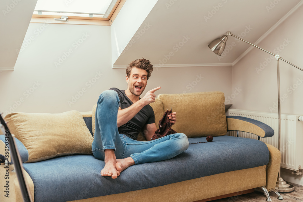 excited young man pointing with finger while knitting on sofa at home excited young man pointing with finger while knitting on sofa at home