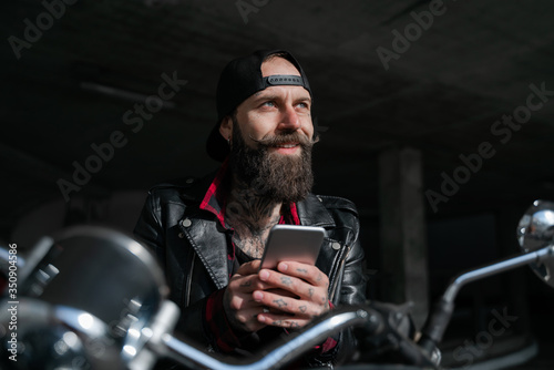 Romantic dreams. Biker traveller, brutal bearded man street racer sitting oh his motorcycle bike and chatting on smartphone with girlfriend. People lifestyle and hobby. Technologies concept.