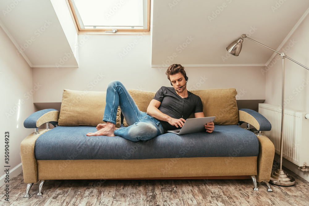 handsome man in headset lying on sofa near laptop while working at home in attic room
