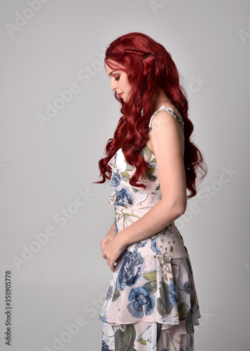 Portrait of a beautiful woman with red hair wearing a flowing floral gown. full length standing pose, isolated against a studio background