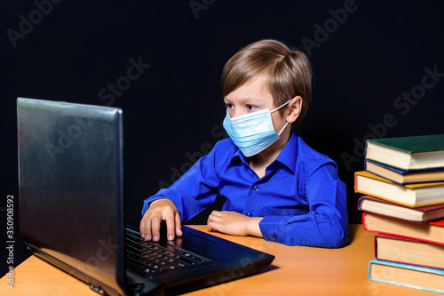 A boy in a blue shirt and a protective mask sits at a Desk and works on a computer. On black background.