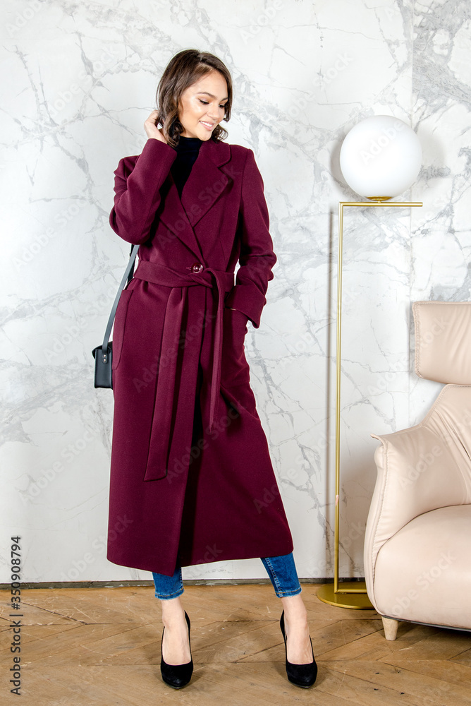 Beauty Fashion Model Girl with curls in burgundy coat in a bright white loft style studio. Beautiful Luxury Winter Woman. Beauty and fashion. Spring and autumn clothes fashion. Girl posing at studio.
