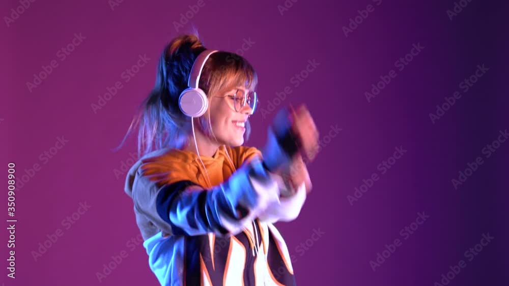 Gen z teen girl model wear headphones listening dj pop music playlist dancing on purple violet neon light color studio background. Stylish 20s hipster fashion woman in hoodie sunglasses at cool party.