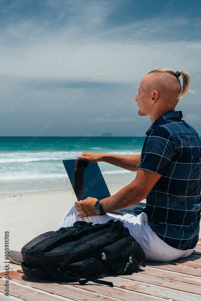 Young successful guy working using a laptop, sitting on the seashore on a sandy beach. Blogger, freelancer makes a content work plan.