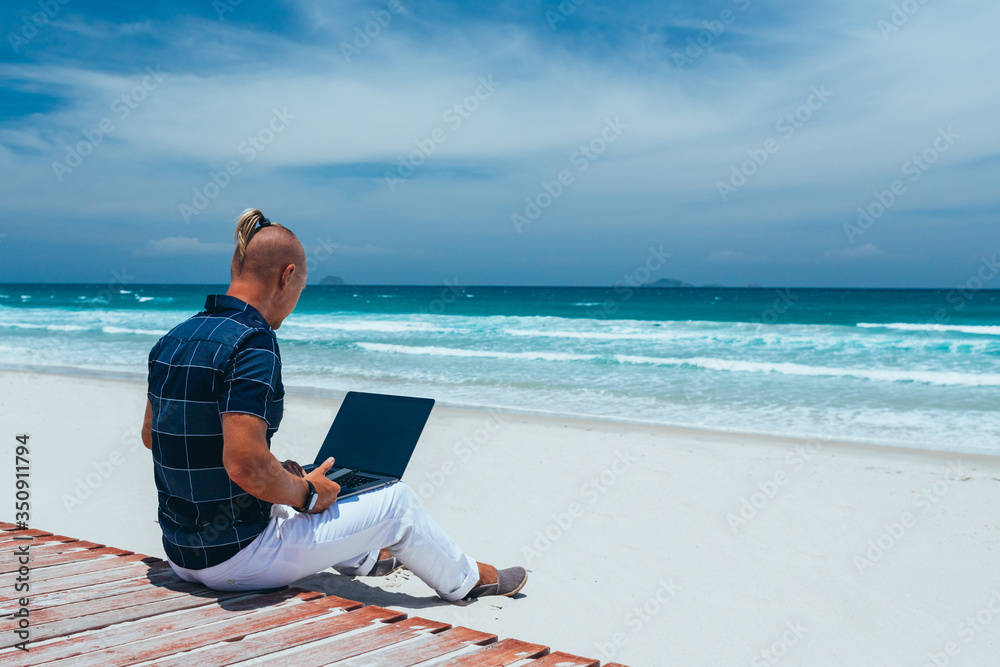 Young successful guy working using a laptop, sitting on the seashore on a sandy beach. Blogger, freelancer makes a content work plan.