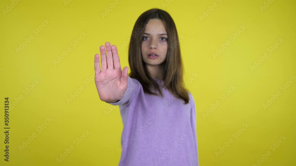 Stop concept: virus, violence, abortion, corruption, inequality, plastic, racism. Serious teenager girl in a pink sweater raises her hand with a palm and says stop on yellow background with copy space Stop concept: virus, violence, abortion, corruption, inequality, plastic, racism. Serious teenager girl in a pink sweater raises her hand with a palm and says stop on yellow background with copy space