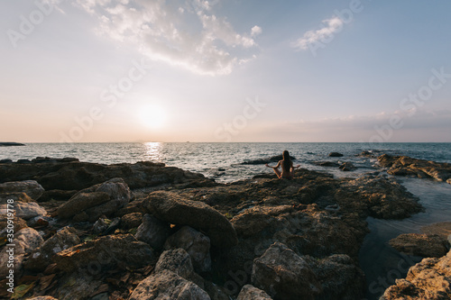 The girl practices yoga on a rocky shore against the background of the sea and sunset sky