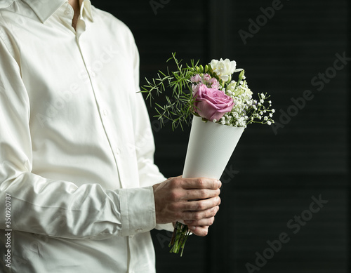 Beautiful bouquet in hands on a background of gray wall. Bouquet of fresh flowers in the package. Male hand holds flowers in paper packaging. Present for woman. Image with selective focus.  photo