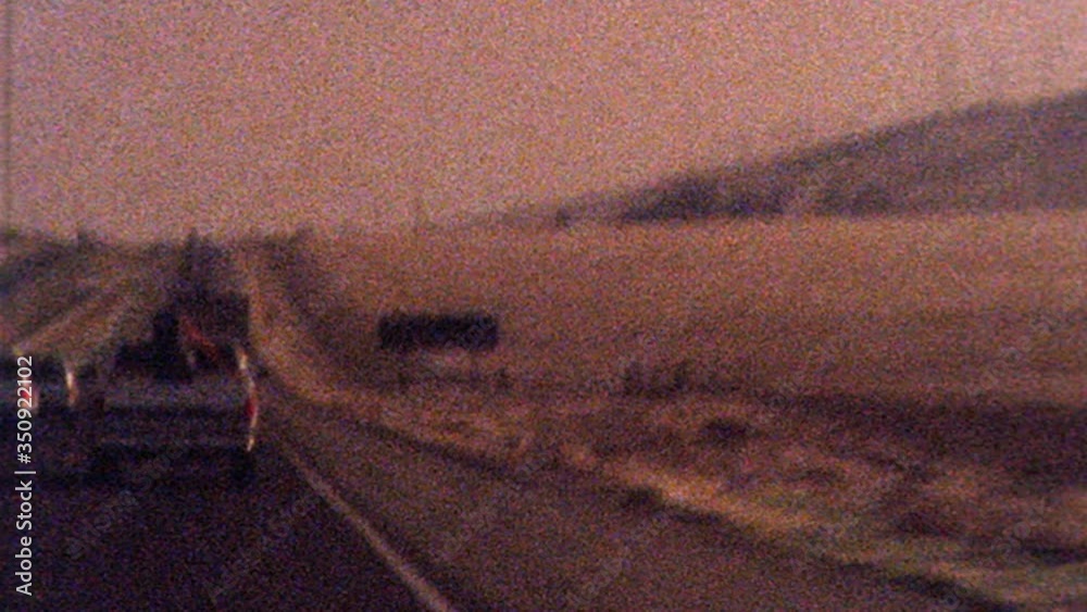 Shaky old looking archival footage drving on 101 highway towards Los Angeles past dessert as sun sets in the distance. Film texture, grit, grain, dust and flash frames create historical look and feel