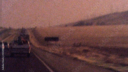 Shaky old looking archival footage drving on 101 highway towards Los Angeles past dessert as sun sets in the distance. Film texture, grit, grain, dust and flash frames create historical look and feel