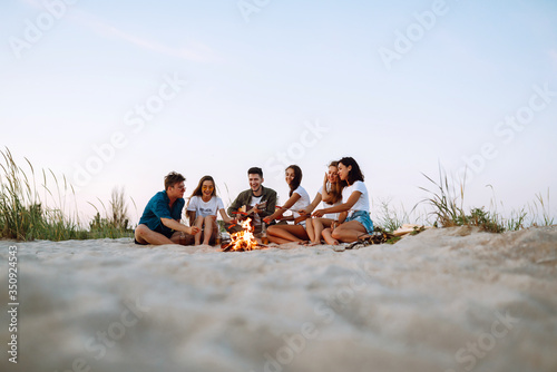 Group of young friends sitting on beach and fry sausages. One man is playing guitar. Summer holidays, vacation, relax and lifestyle consept. Camping time.
