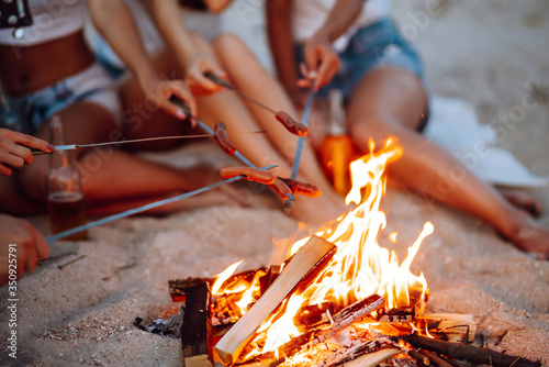 Fried sausages on bonfire. Group of young friends sitting on beach and fry sausages. Summer holidays, vacation, relax and lifestyle consept. Camping time.