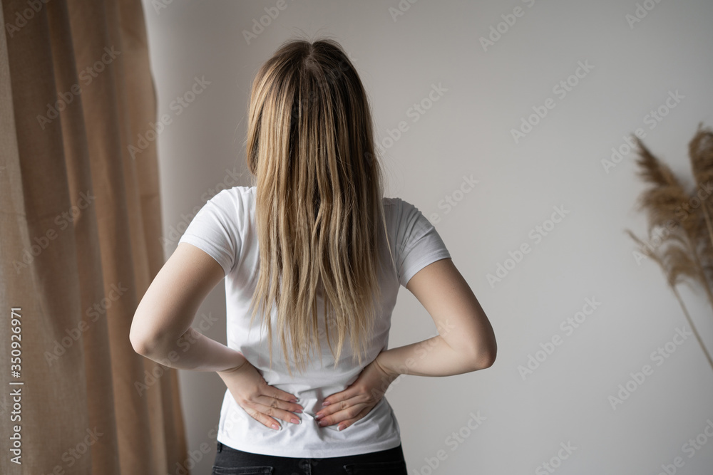 rear view of a young woman standing in a room and suffering from back pain.