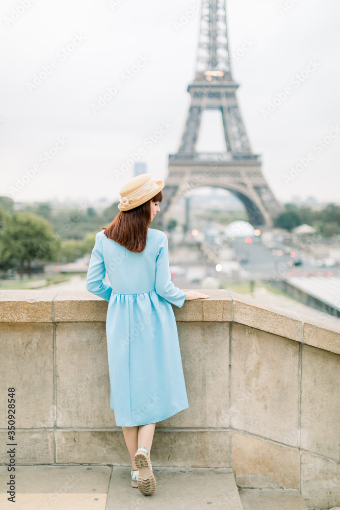 Obraz premium Pretty young caucasian girl walking near stairs, Eiffel Tower in background, Paris. Charming lady in blue dress staying back to camera and looking at the beautiful view of Paris