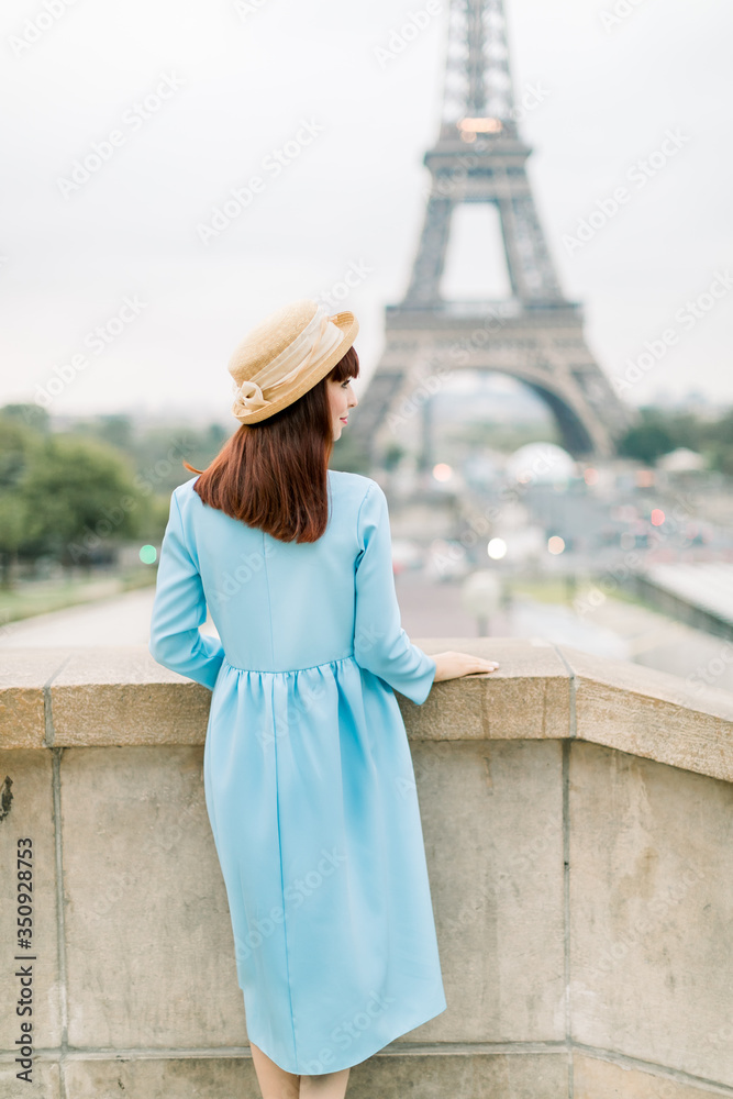 Back view of stylish tourist woman in blue dress and hat sightseeing Eiffel Tower in Paris, France