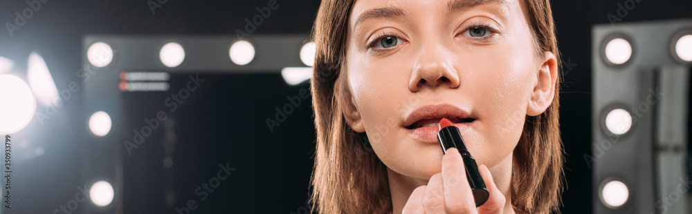 Panoramic orientation of beautiful woman applying red lipstick and looking at camera in photo studio