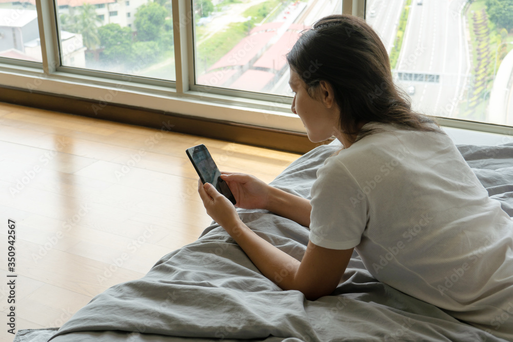 A beautiful young brunette girl is texting on the phone while lying on a bed by a panoramic window with a beautiful view from a high floor.