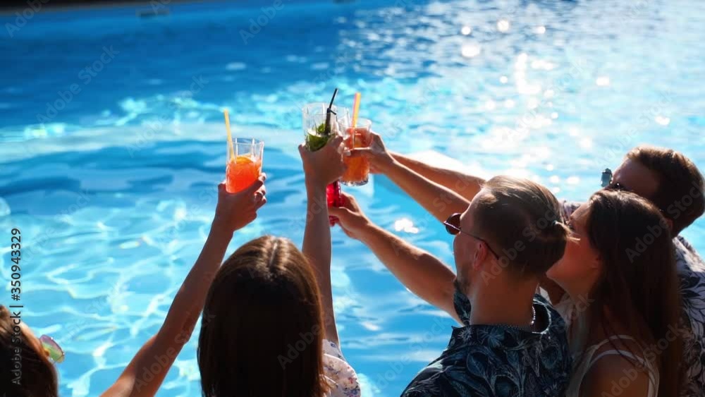 Group of friends having fun at poolside party clinking glasses with fresh cocktails sitting by swimming pool on sunny summer day. People toast drinking beverages at luxury villa on tropical vacation.