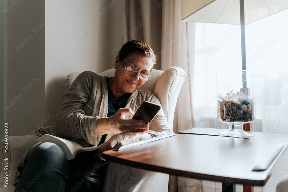 An older woman is sitting on the sofa with a book. She is typing on her phone while smiling. She is protecting herself and staying home. Pandemic concept. An older woman is sitting on the sofa with a book. She is typing on her phone while smiling. She is protecting herself and staying home. Pandemic concept.