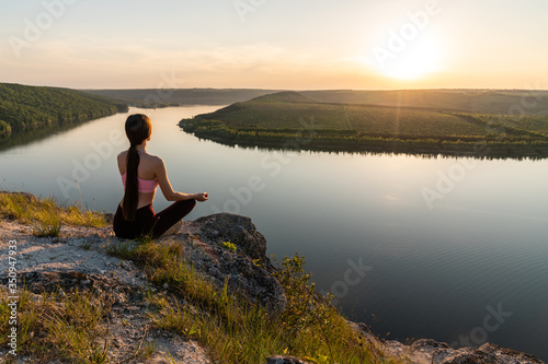 Woman in pink top and black leggings is practicing yoga performing yoga-lotus position outdoors, near a riverside
