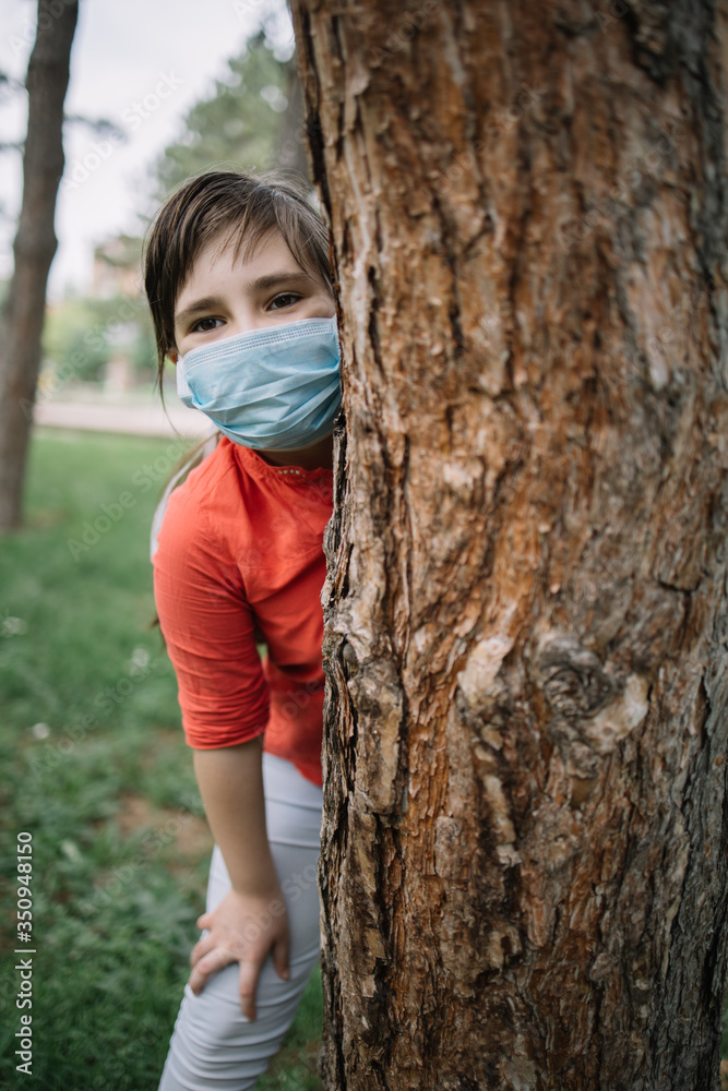 Happy child wearing antivirus mask while standing behind tree. Cute little child wearing medical mask while hiding behind tree in park.