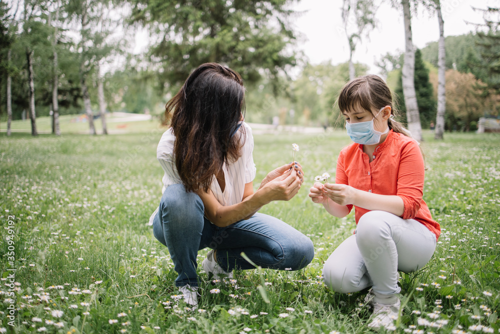 Girls with antivirus masks sitting on lawn in park and gathering flowers. Woman and kid with protection masks squatting on meadow with camomile during pandemic.