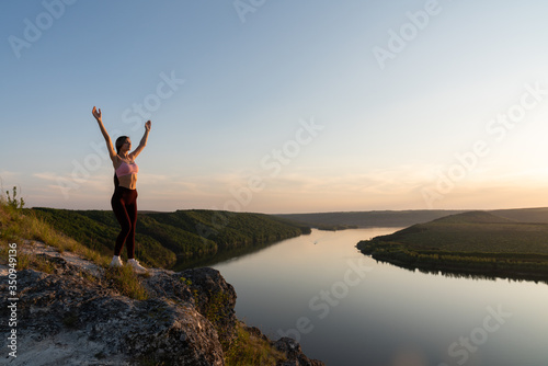 Young female hiker in sportswear stands with raised hands on the rock on the hill