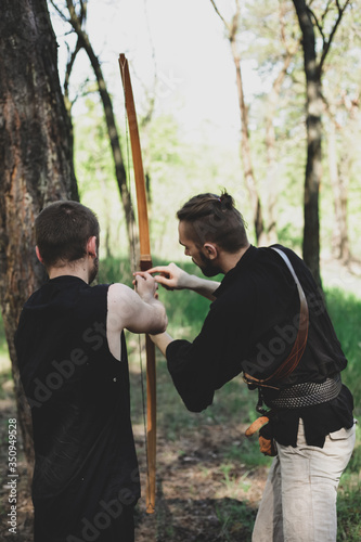 A man teaches to shoot with a bow. Two men, one teacher. Archery in nature. The boy holds a bow in his hands.
