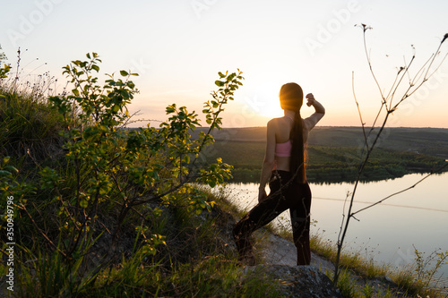 Back view of young joyful woman standing at the top with stretched arm, expressing happy emotions, enjoying beautiful landscape, looking at colorful sunrise