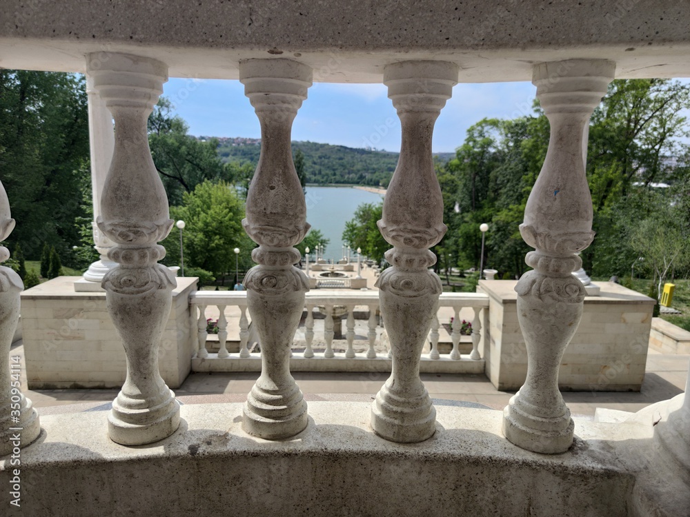 Fototapeta premium Stone stairs to the lake. Around the park with green trees and firs. Blue sky with clouds. View through the white columns.