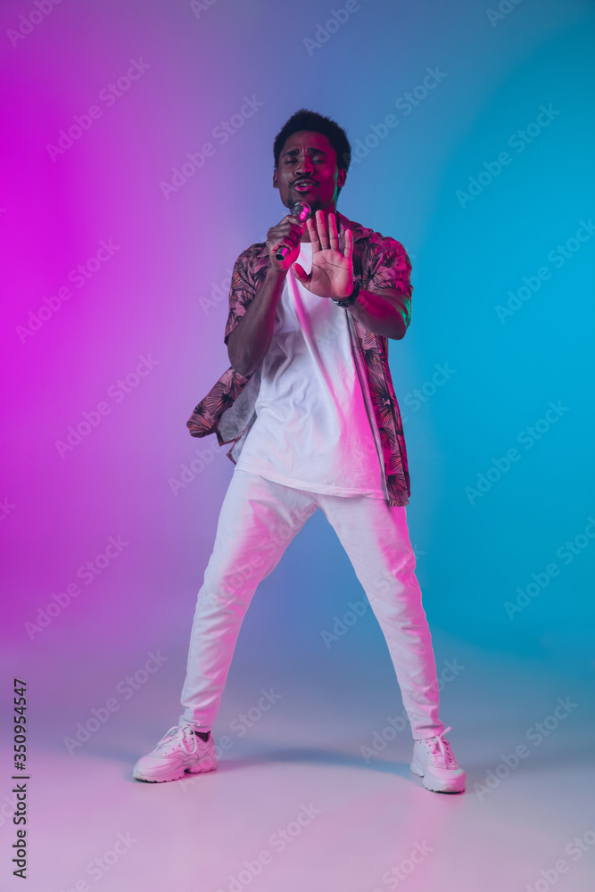 African-american male singer portrait isolated on gradient studio background in neon light. Beautiful male model with microphone. Concept of human emotions, facial expression, ad, music, art. African-american male singer portrait isolated on gradient studio background in neon light. Beautiful male model with microphone. Concept of human emotions, facial expression, ad, music, art.