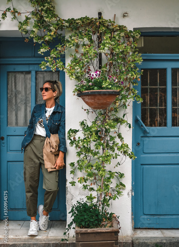 Pretty fashionable woman standing in doorway of old house. Cool dressed lady in front of turquoise wooden door with flowers around. photo