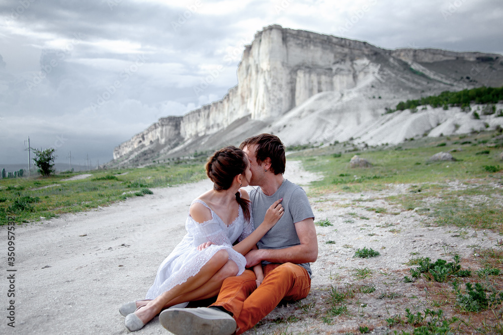 Happy couple in relationship walking in the beautiful place. Female dressed white dress and posing against the white mountain cloudy sky. Man hugging his girlfriend