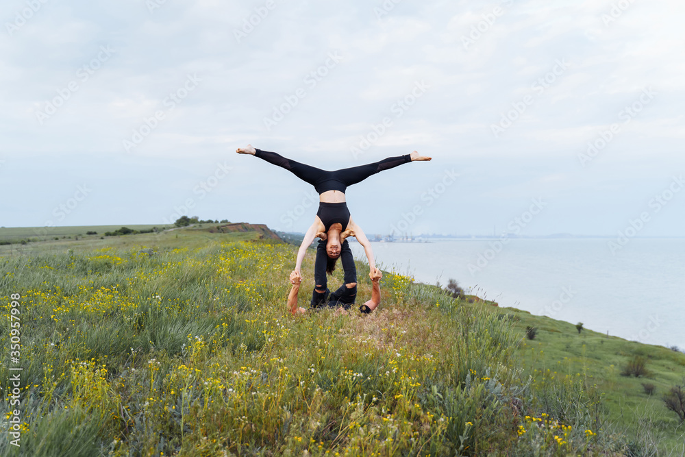 Attractive couple practicing yoga outdoors on the shore of the sea. Girl doing splits, being upside down during acroyoga, lying on the legs of a man. Sport exercises on the hill. Long distance shot Attractive couple practicing yoga outdoors on the shore of the sea. Girl doing splits, being upside down during acroyoga, lying on the legs of a man. Sport exercises on the hill. Long distance shot