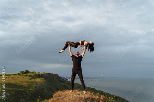 Full-length portrait. Attractive muscular, pumped-up young man is doing in acroyoga with his girlfriend. Instead of a bar, he lifts her up on outstretched arms and smiles. Standing on the edge of hill © Daria
