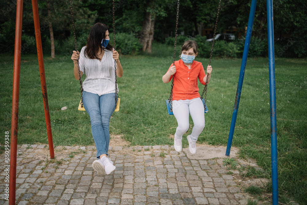 Brunette mother and daughter playing in the park during pandemic. Pretty girl and woman wearing protection mask while enjoying playground facilities.