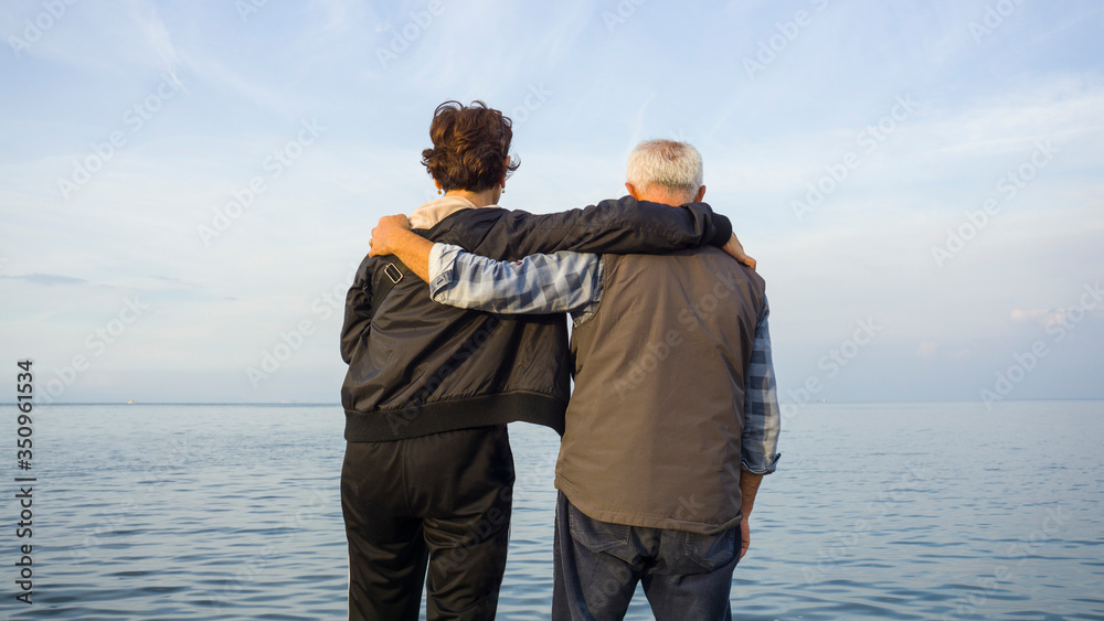 Beautiful old couple putting their arms around shoulders, watching seascape on shore. Romantic senior man & woman enjoying now, today. Seize the day moment. Photo of two 60s person together. Beautiful old couple putting their arms around shoulders, watching seascape on shore. Romantic senior man & woman enjoying now, today. Seize the day moment. Photo of two 60s person together.