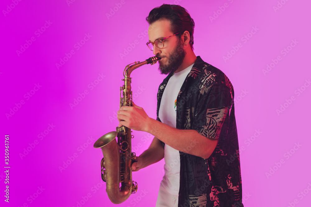 Fototapeta premium Young caucasian jazz musician playing the saxophone on gradient pink studio background in neon light. Concept of music, hobby, festival. Joyful, cheerful attractive guy. Colorful portrait of artist.