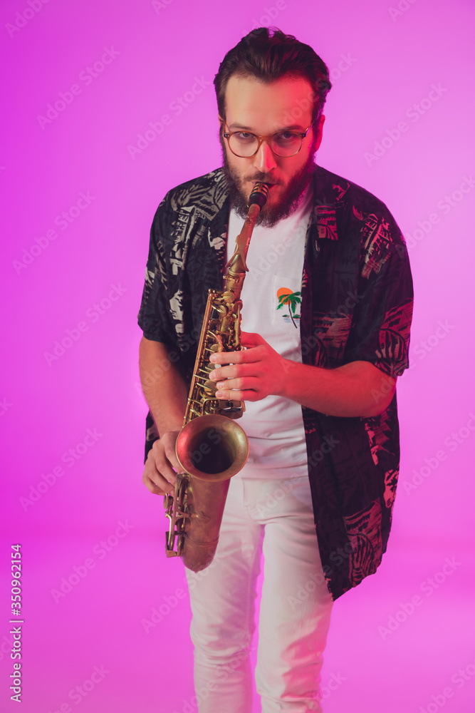 Fototapeta premium Young caucasian jazz musician playing the saxophone on gradient pink studio background in neon light. Concept of music, hobby, festival. Joyful, cheerful attractive guy. Colorful portrait of artist.