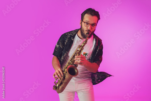Photography Young caucasian jazz musician playing the saxophone on gradient pink studio background in neon light