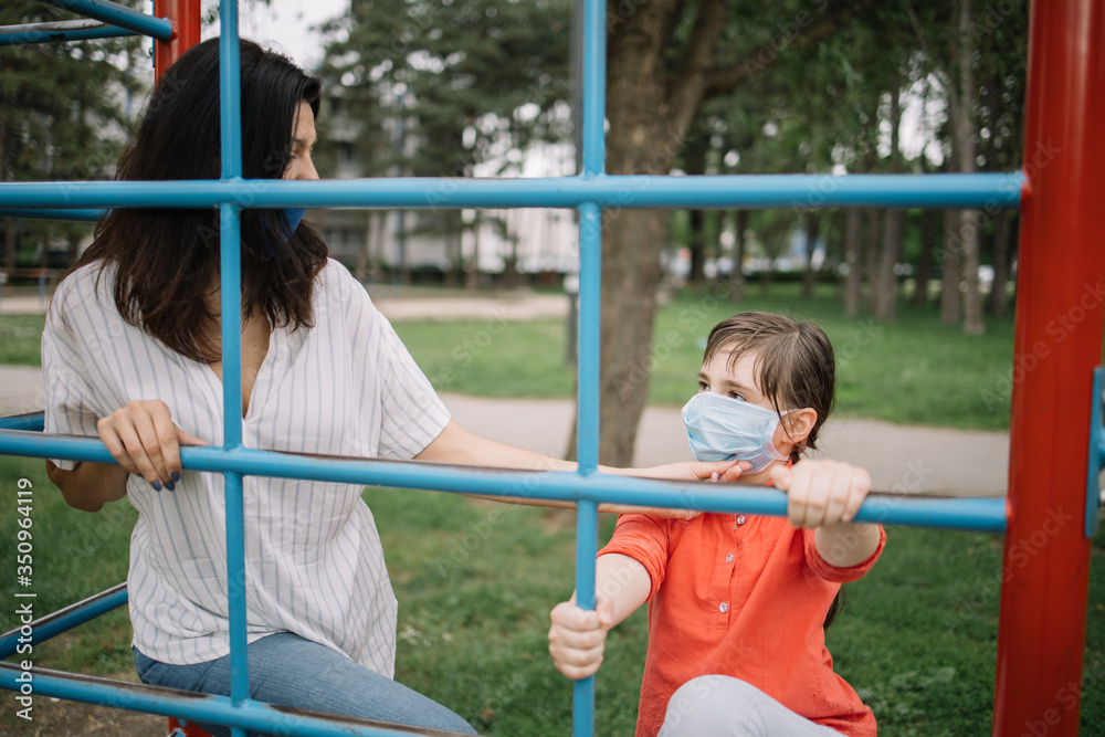 Obraz premium Careful woman fixing position of medical mask on girl's face at playground. Mother and daughter protecting their faces with antivirus masks while playing in the park.