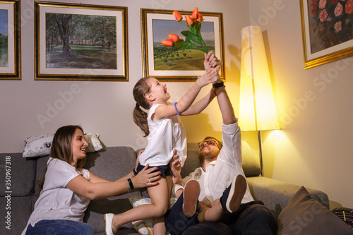 famiglia di quattro persone gioca allegramente nel divano e la figlia piccolina prende il fiore che il papà vuole dare alla mamma photo