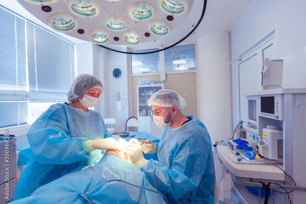 Male surgeon performs surgical procedures in the operating room, while his assistant monitors the patient condition Male surgeon performs surgical procedures in the operating room, while his assistant monitors the patient condition