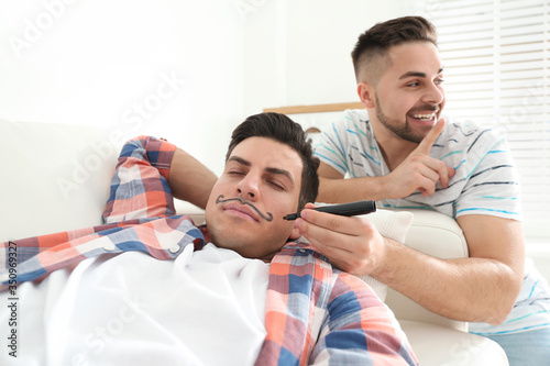 Young man drawing mustache on face of sleeping friend indoors. April fool's day photo