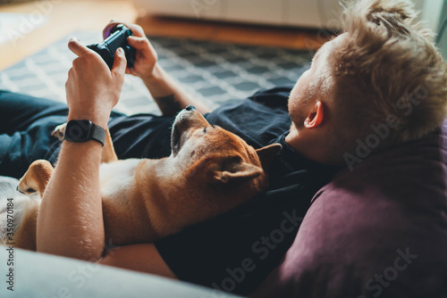 Young hipster guy playing video games at home holding game joystick relaxing on couch with his best friend dog, Home Leisure Concept photo