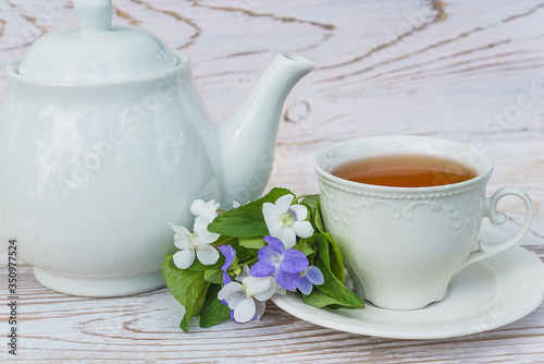 Tea time concept in retro style. White cup of tea, tea pot and bouquet of white and blue violets on white paint rustic wooden background. Close up