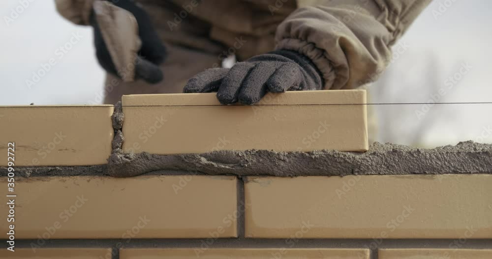 Close-up of construction work, male bricklayer puts another yellow brick into mortar, neatly smooths edges with trowel, taps, works well, professional, building private house from brick, construction