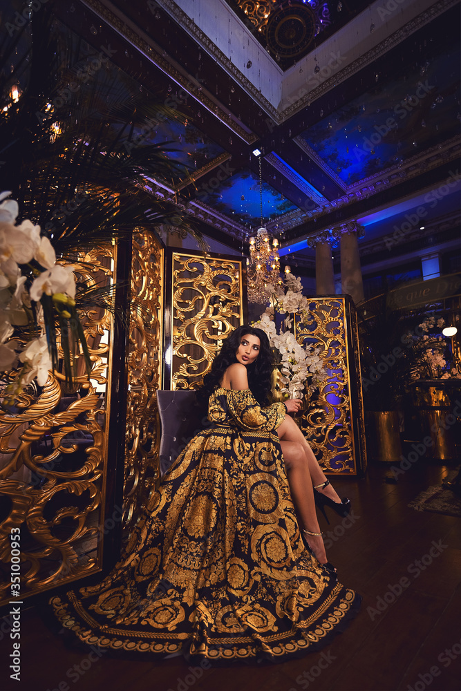 Portrait of a beautiful tanned brunette girl in a silk black and gold dress in a luxurious interior with gold decor, crystal and orchids
